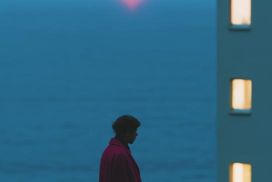A solitary woman in a red coat gazes at the ocean during a serene sunset, near a lit building