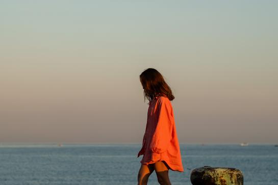 A girl, in a long pink shirt, greets the morning on a pier in the ocean, thinking about life