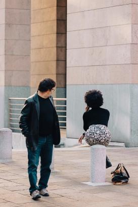 Attractive woman sitting on a pole in the city is talking on the phone and looking away. There is a man walking next to the girl and he turns around to look at her