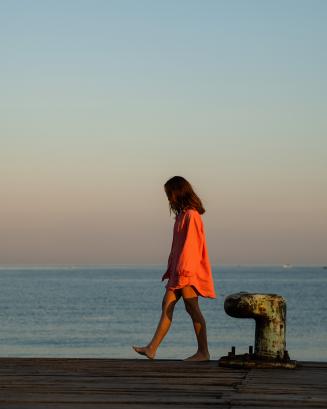 A girl, in a long pink shirt, greets the morning on a pier in the ocean, thinking about life